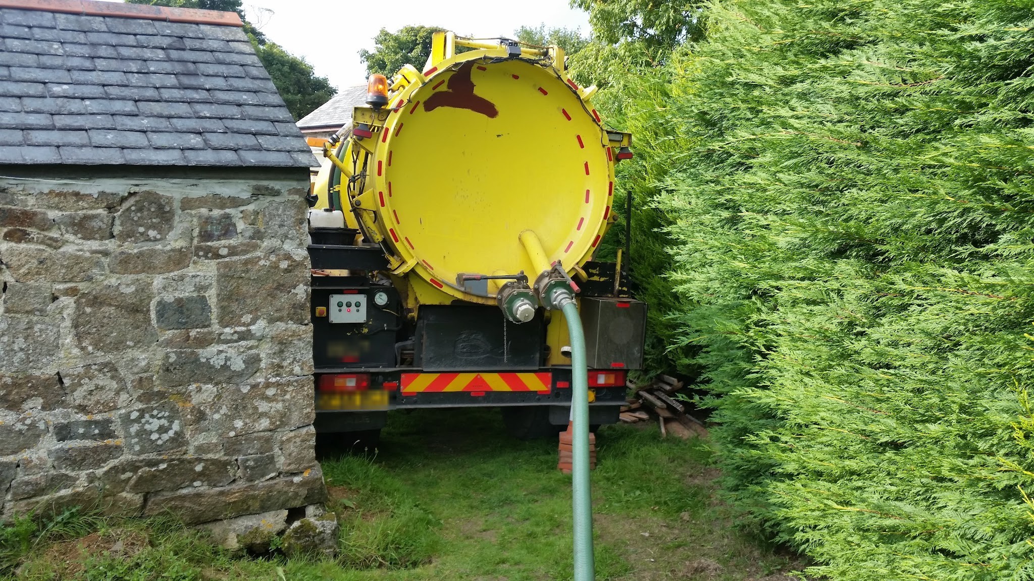 The yellow tanker reversed down a hedged lane beside a stone outbuilding, hose out