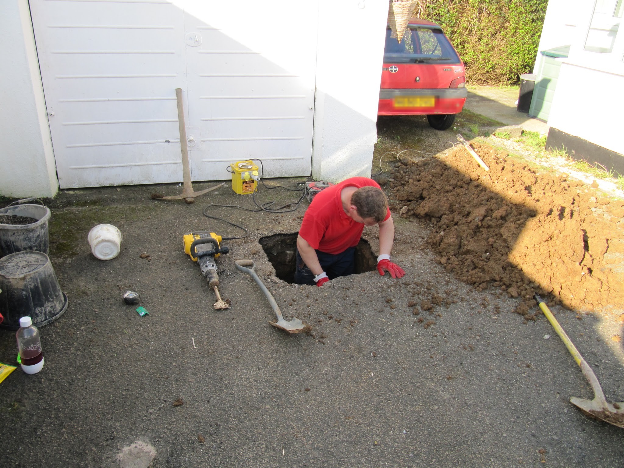 An engineer in a red top down a small excavation beside a garage, with tools laid out