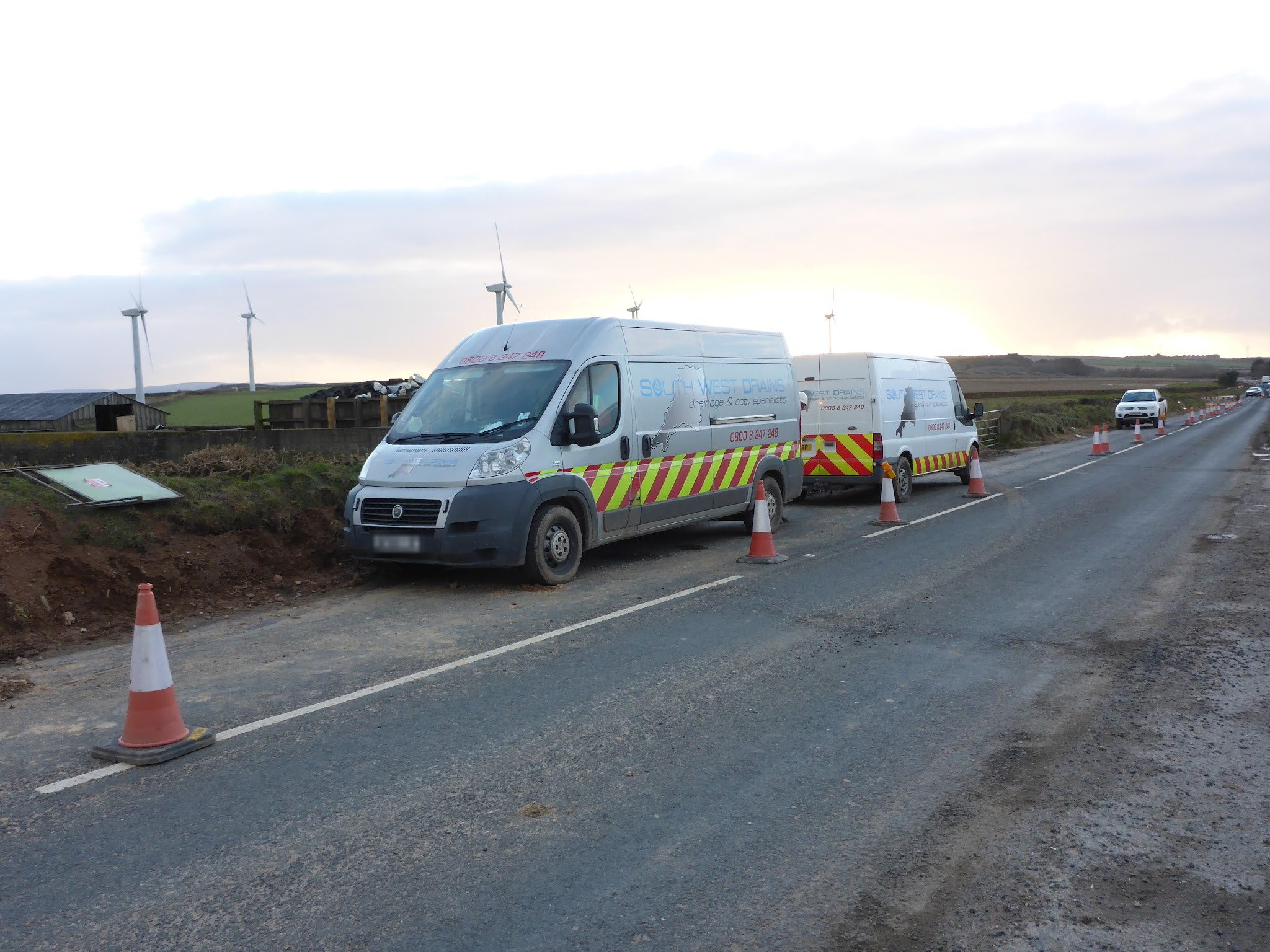Two South West Drains vans parked by a coned-off road near wind turbines