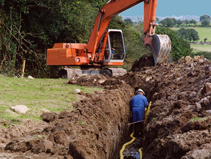 A digger opening a long trench across a field with a worker in the trench