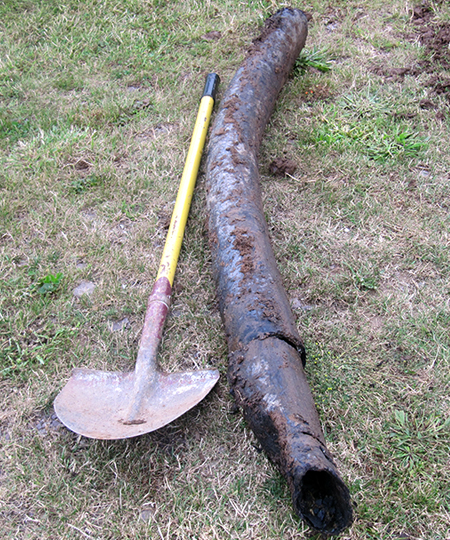 A failed length of pitch fibre pipe lying on grass beside a spade
